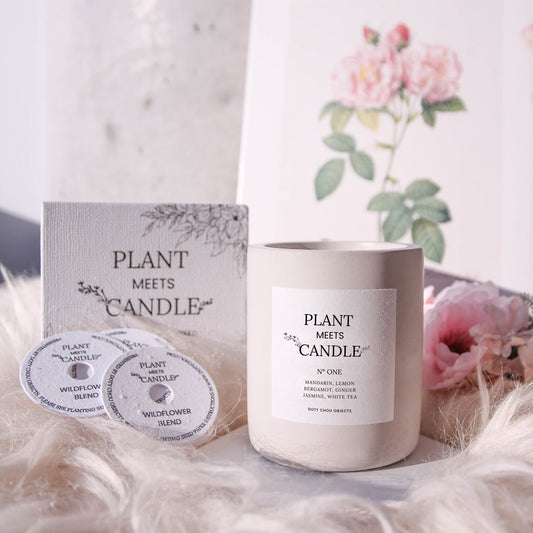 A light grey concrete vessel containing Plant Meets Candle No. One is displayed in a floral background alongside its product box and seed paper dustcover.