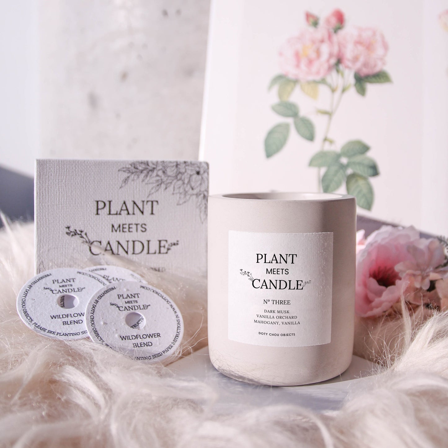 A light grey concrete vessel containing Plant Meets Candle No. Three is displayed on a floral background alongside its product box and seed paper dustcover.